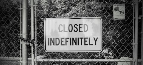A chain-link gate in black and white with a sign that says closed indefinitely, and a smaller warning with gruesome icons for entrapment (a person being smashed) and pinching (a hand going through gears)
